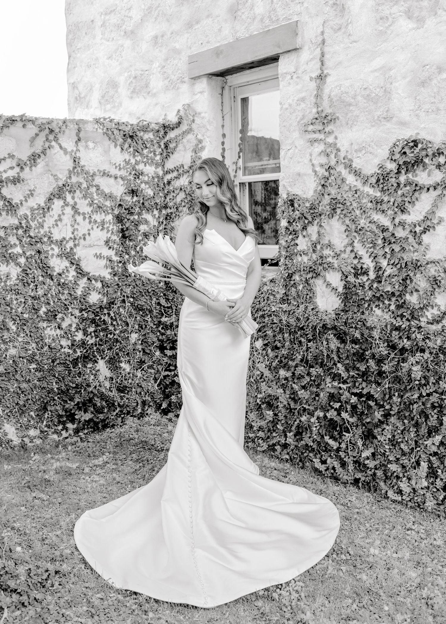 Black and white bridal portrait against ivy-covered wall showing elegant fitted wedding gown with long train.