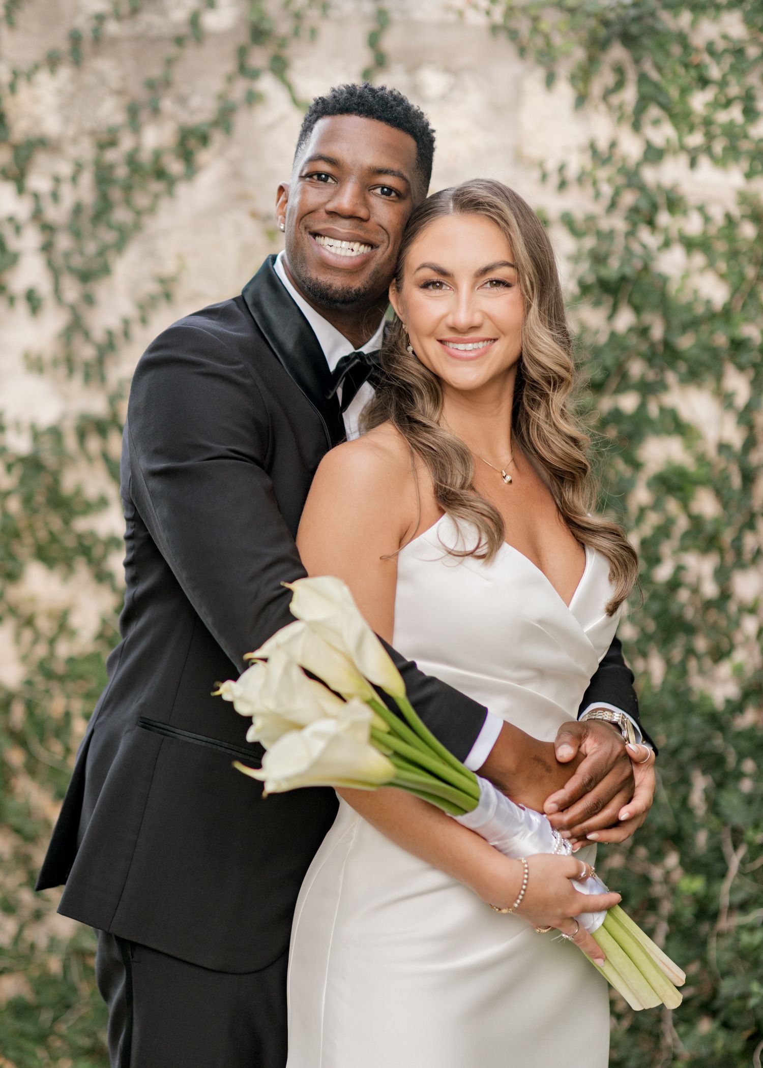 Romantic wedding portrait of couple embracing while holding white calla lily bouquet against ivy wall.