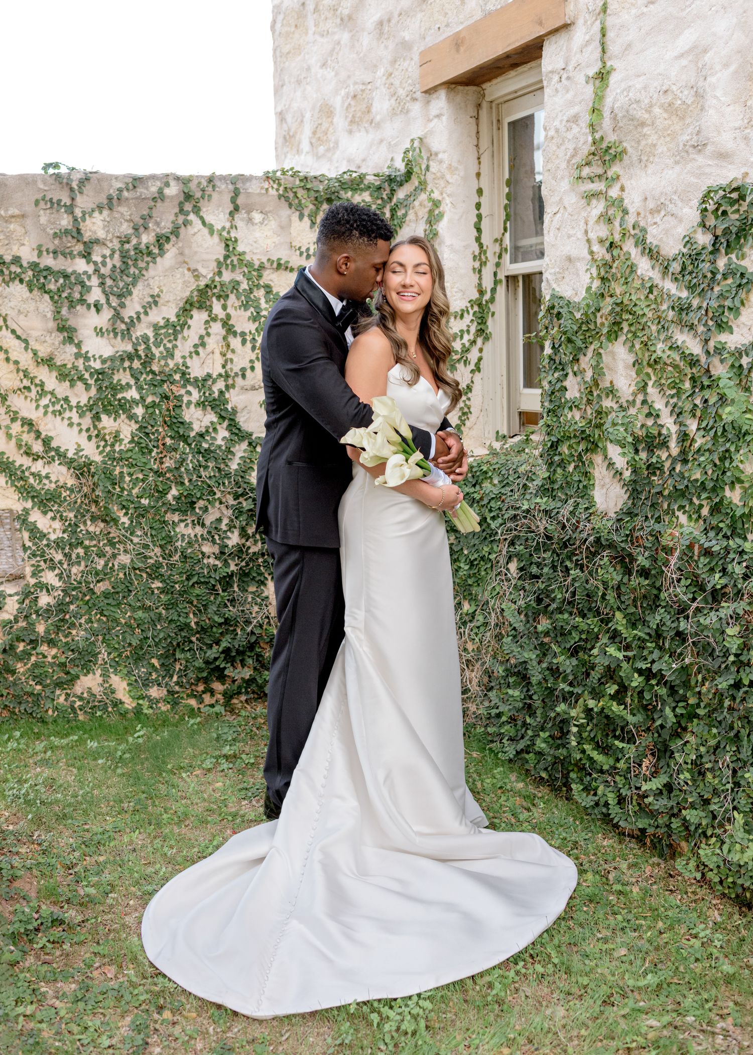 Romantic full-length wedding portrait of couple embracing against stone wall with climbing vines.