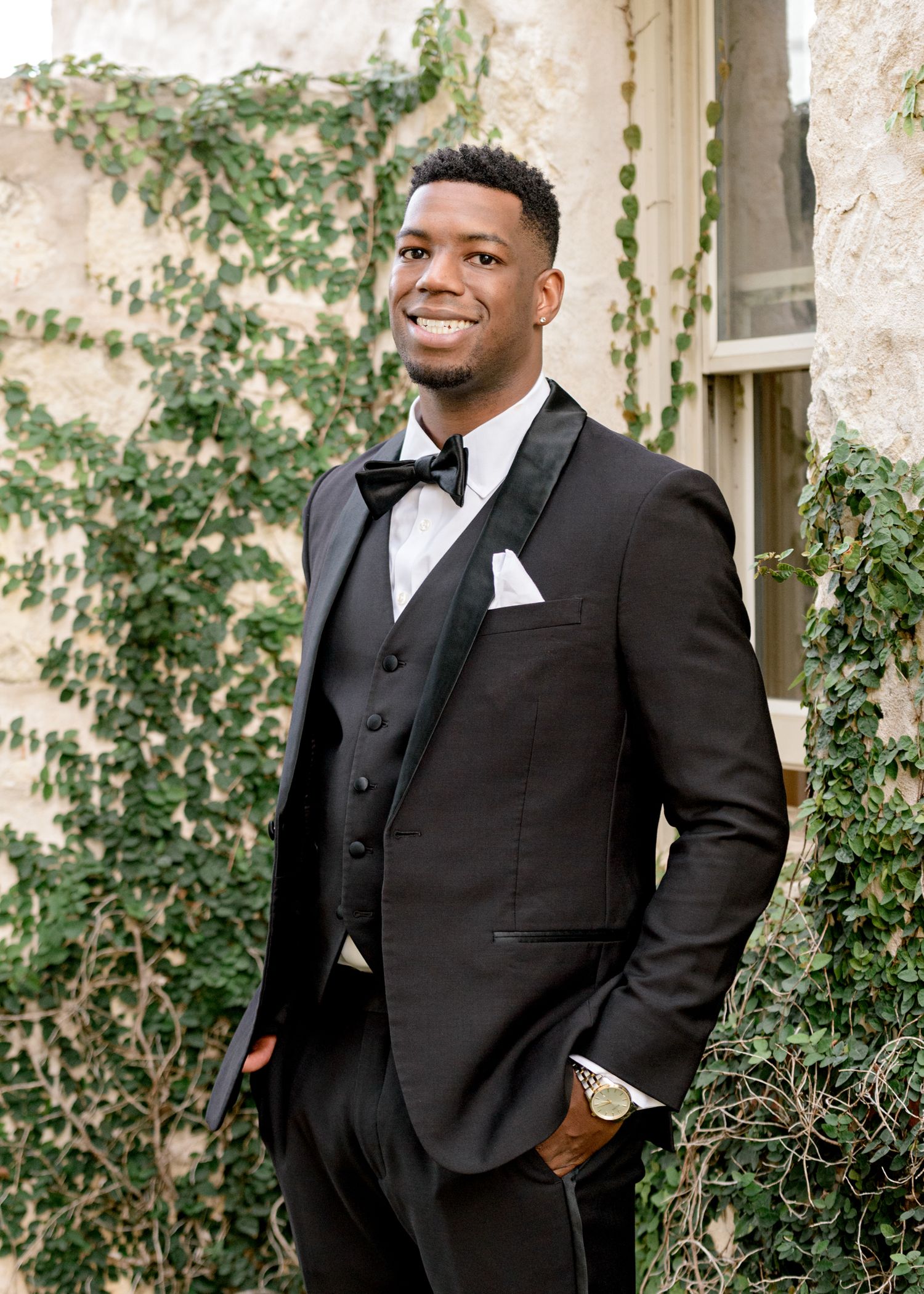 Groom in three-piece black tuxedo poses confidently against ivy-covered stone wall.