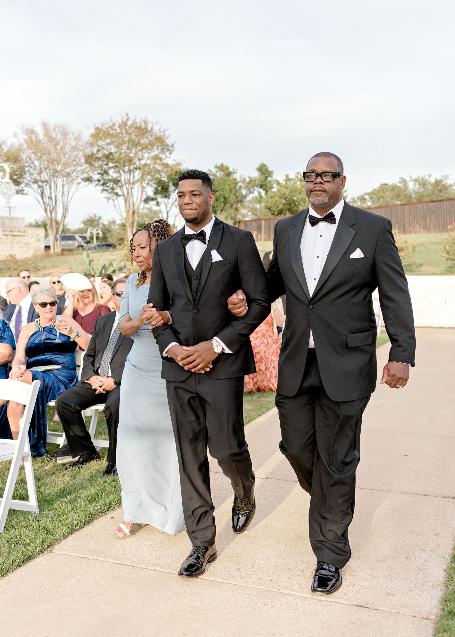A wedding processional on a paved path with people in formal black tuxedos walking past seated guests.