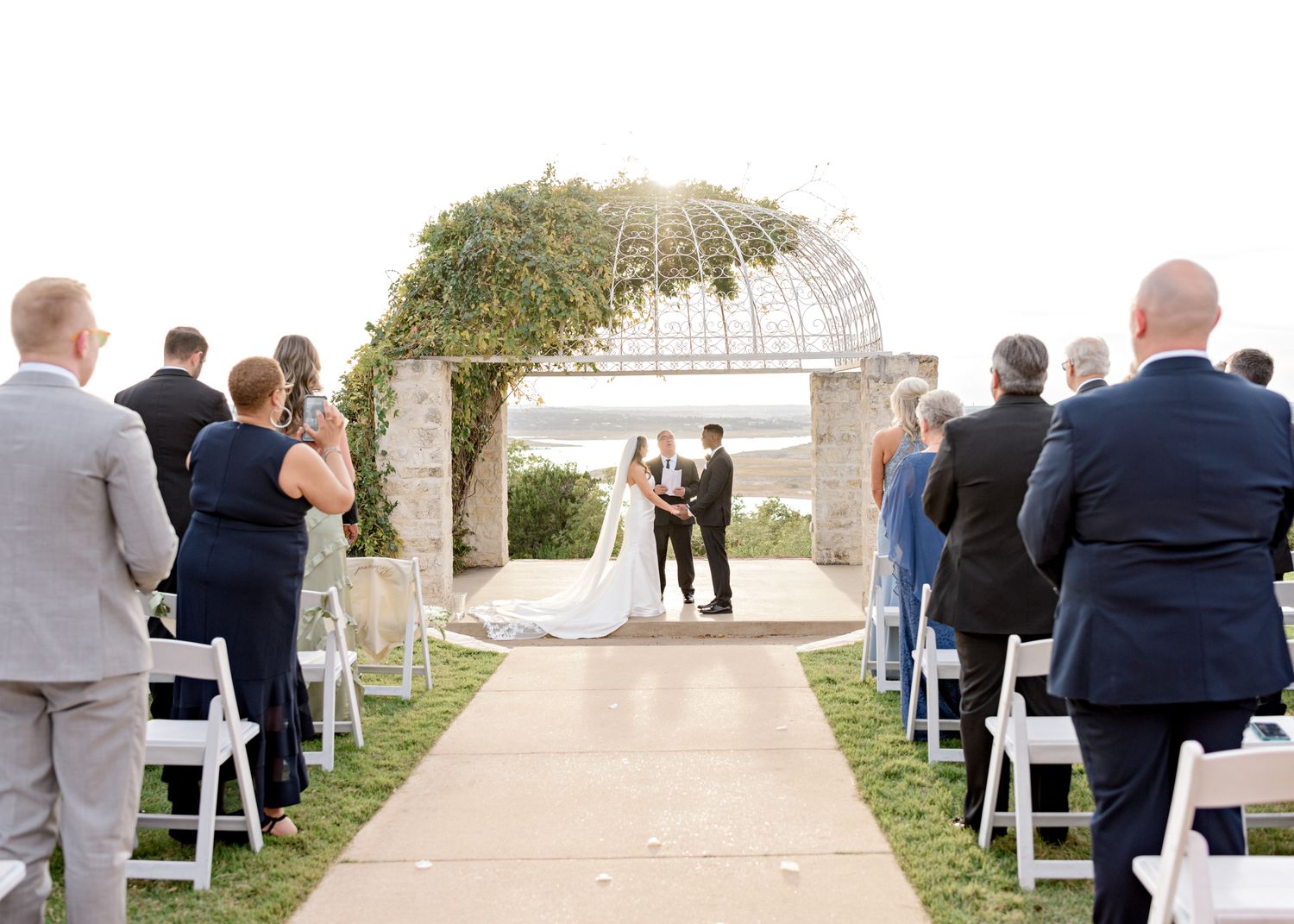 Wedding ceremony under an elegant vine-covered arch with guests seated on white chairs overlooking water.