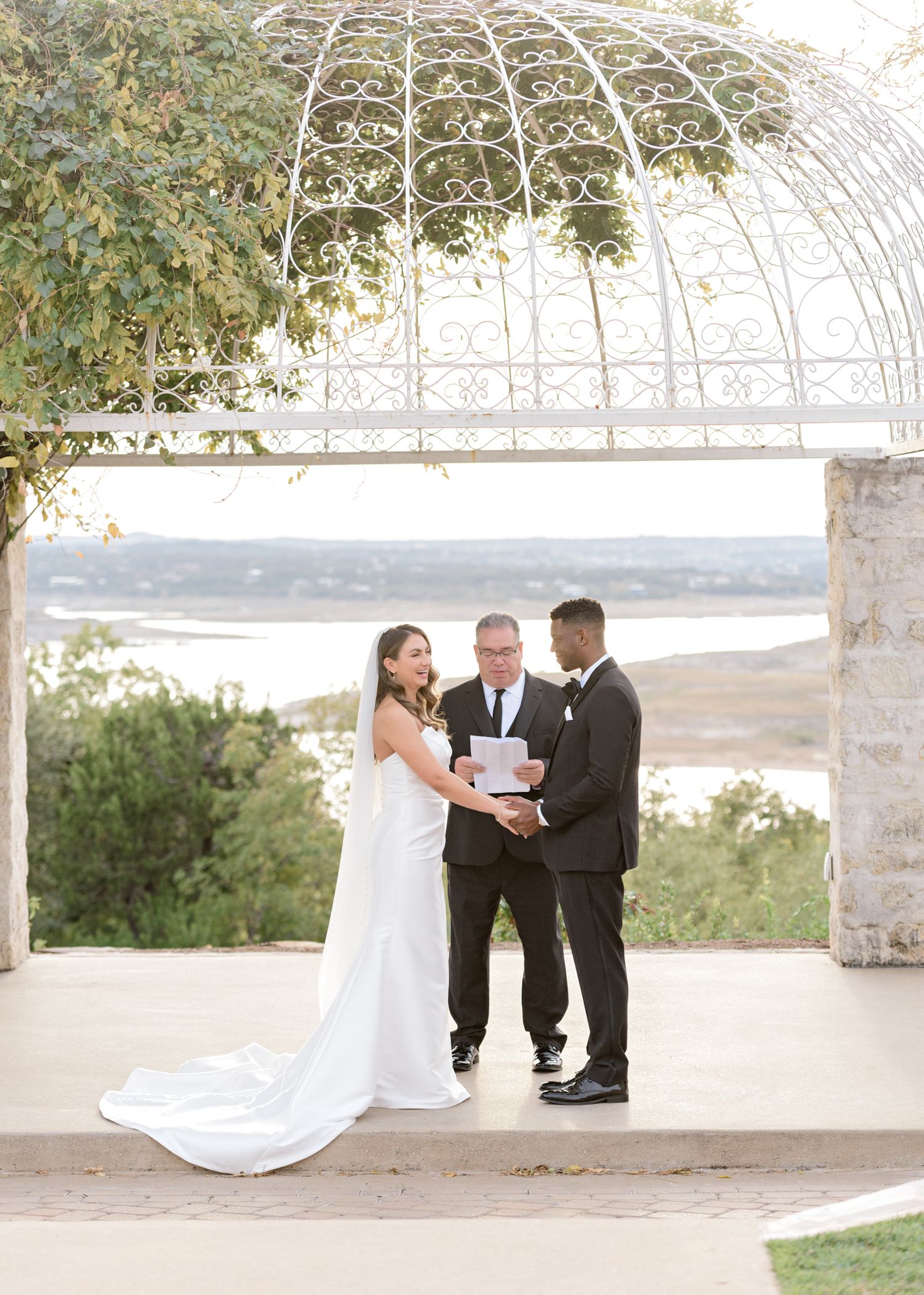 Wedding ceremony under an ornate white metal dome with waterfront views and natural greenery.