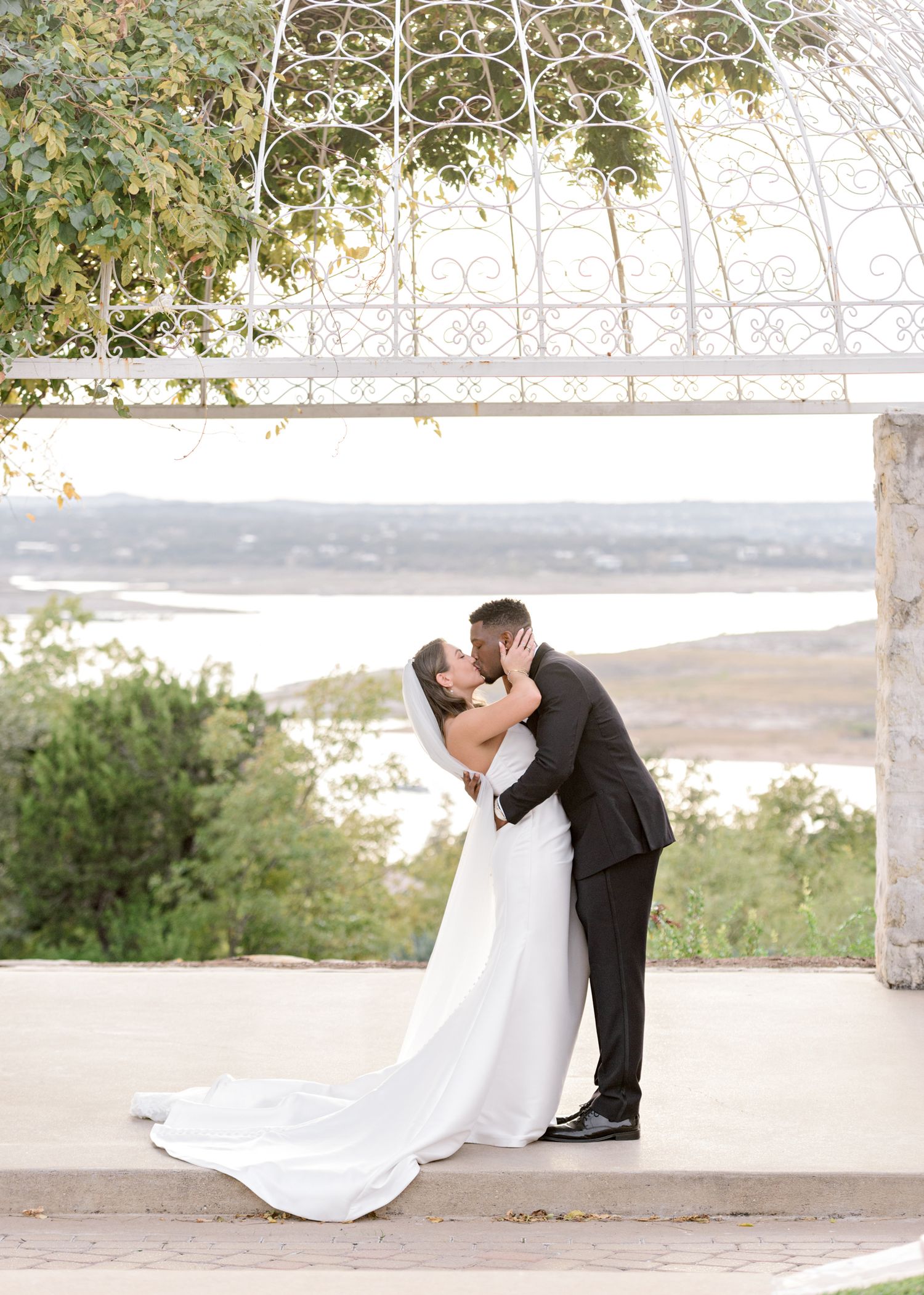 A romantic wedding kiss under an ornate gazebo overlooking a scenic waterfront view.