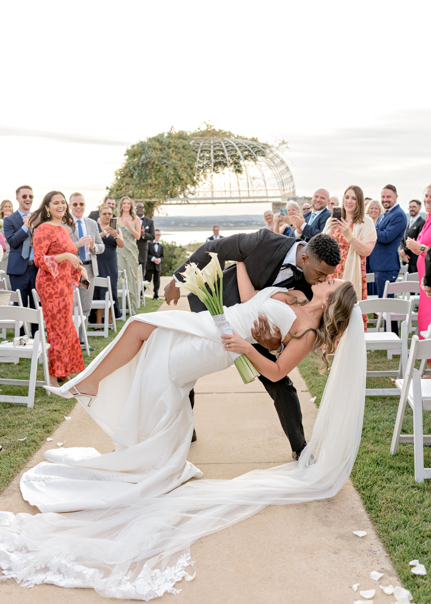 A dramatic wedding dip kiss on a lawn with guests cheering in the background.