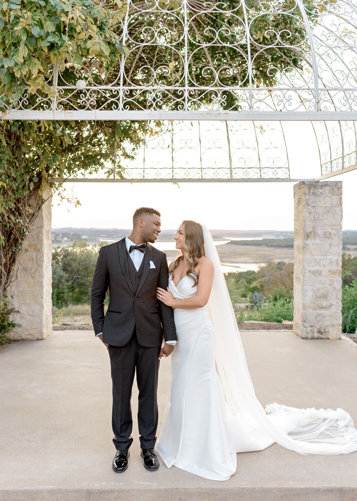 Elegant wedding couple poses under an ornate garden pergola with scenic backdrop.