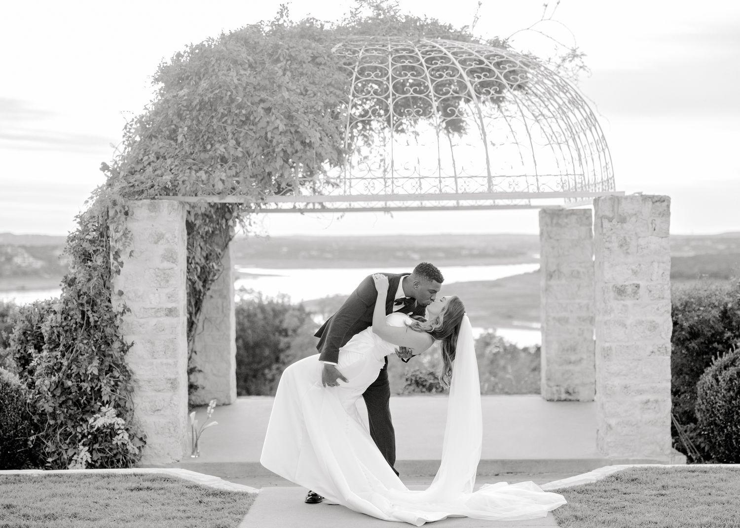 Dramatic black and white wedding photo with couple kissing under arched garden structure.