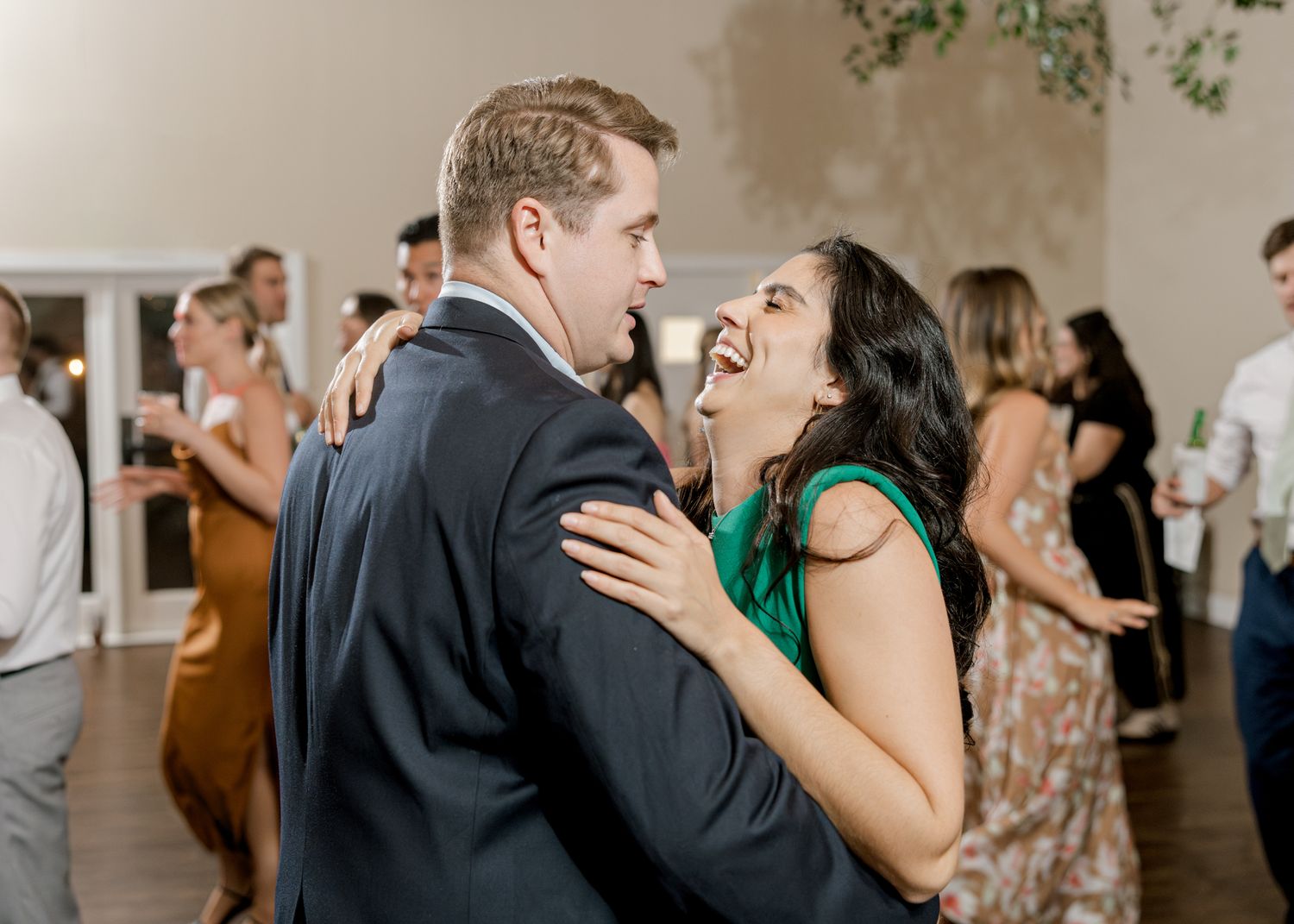 Joyful couple share a laugh while dancing together at an indoor wedding reception under greenery decorations.