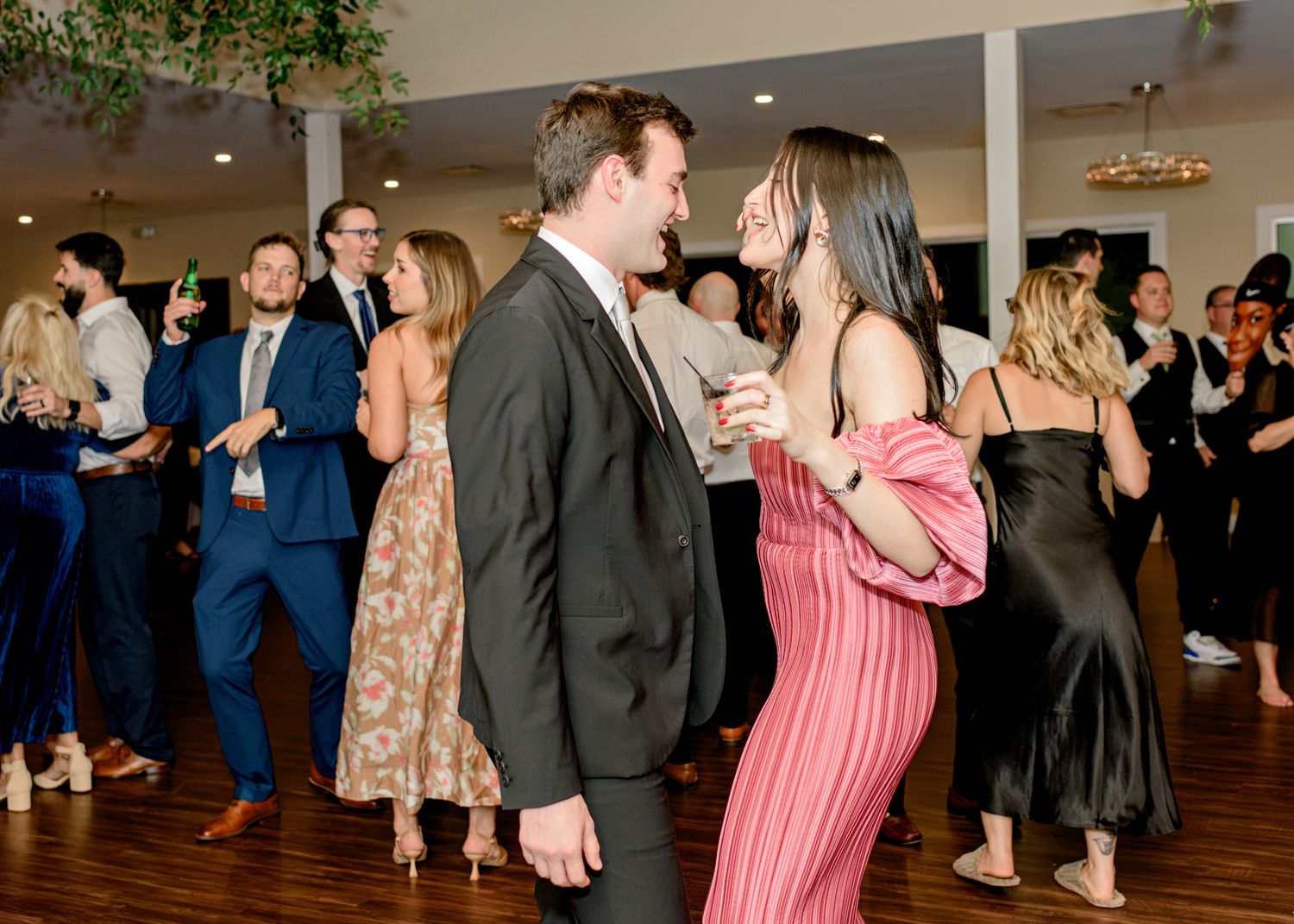 Wedding guests dancing and socializing on a wooden dance floor under decorative greenery.