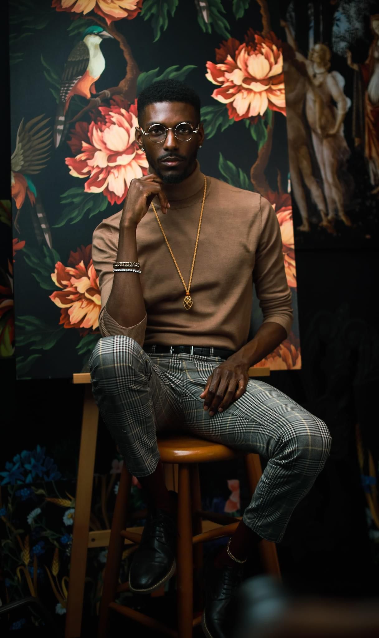 Fashionable person in brown sweater and plaid pants seated on stool against floral wallpaper.