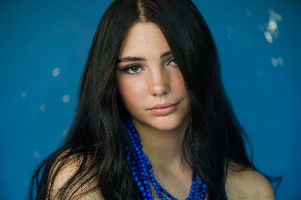 Person with long dark hair wearing bright blue beaded necklace against turquoise background.