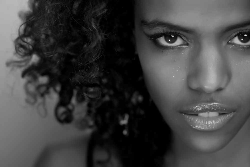 Black and white close-up portrait featuring curly hair, intense gaze, and glossy lips against a dark background.