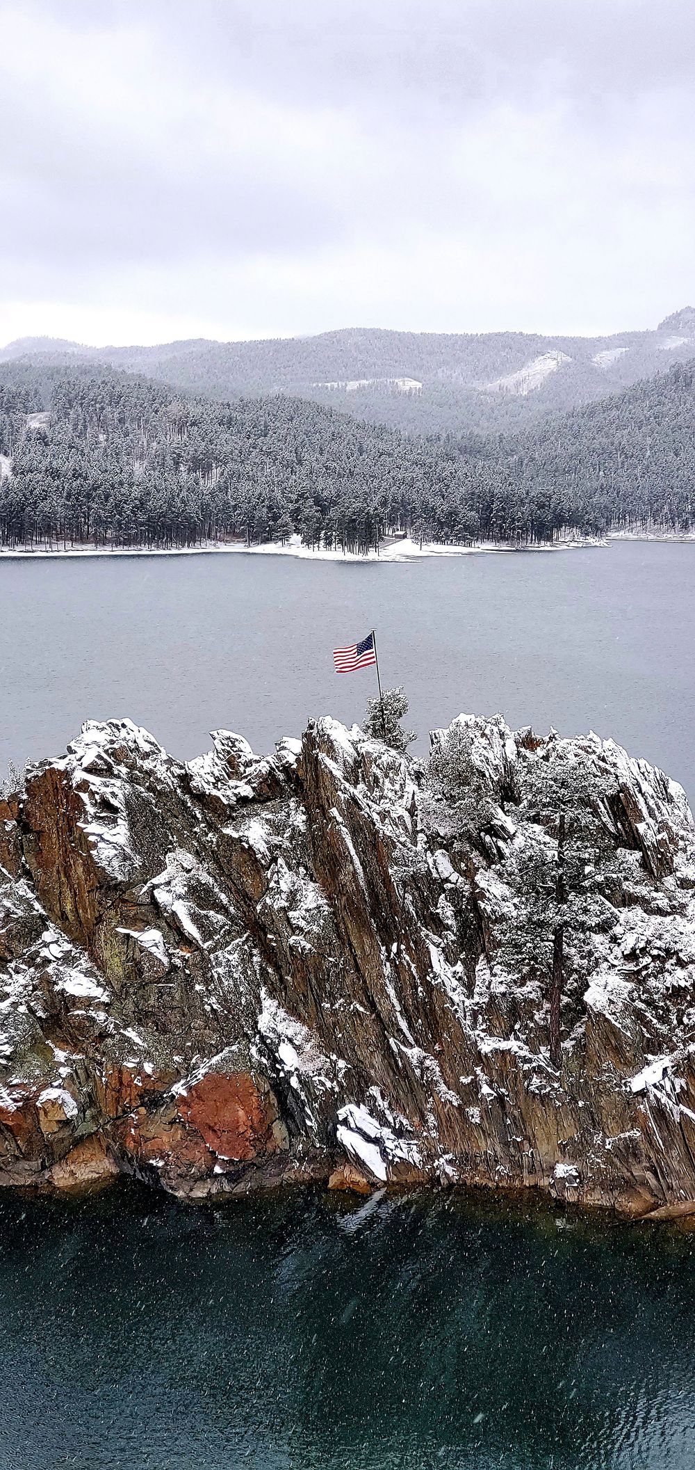 Snow-dusted rocky cliffs rise from dark waters with evergreen-covered mountains and an American flag visible in the distance.