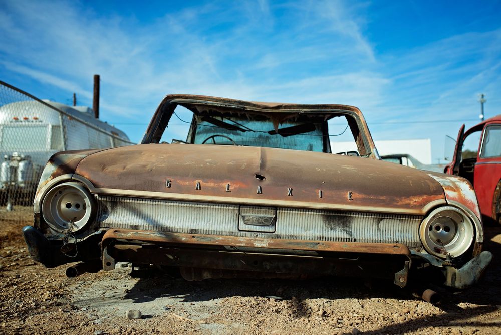 Abandoned vintage Ford car rusting in a junkyard under blue sky.