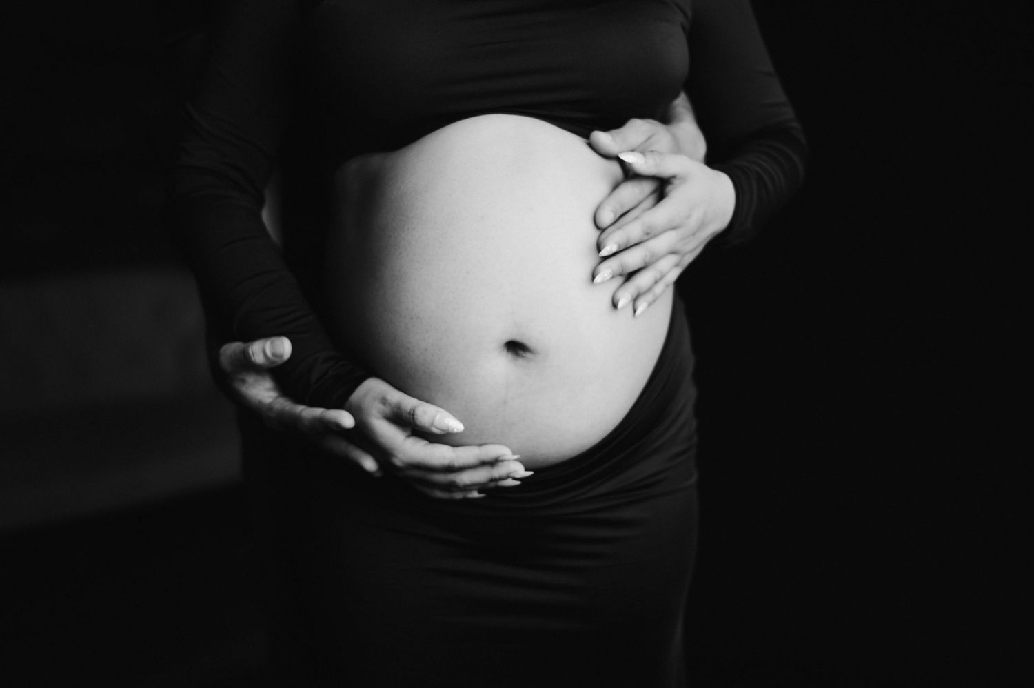Black and white artistic maternity photo showing hands cradling a pregnant belly against dark background.