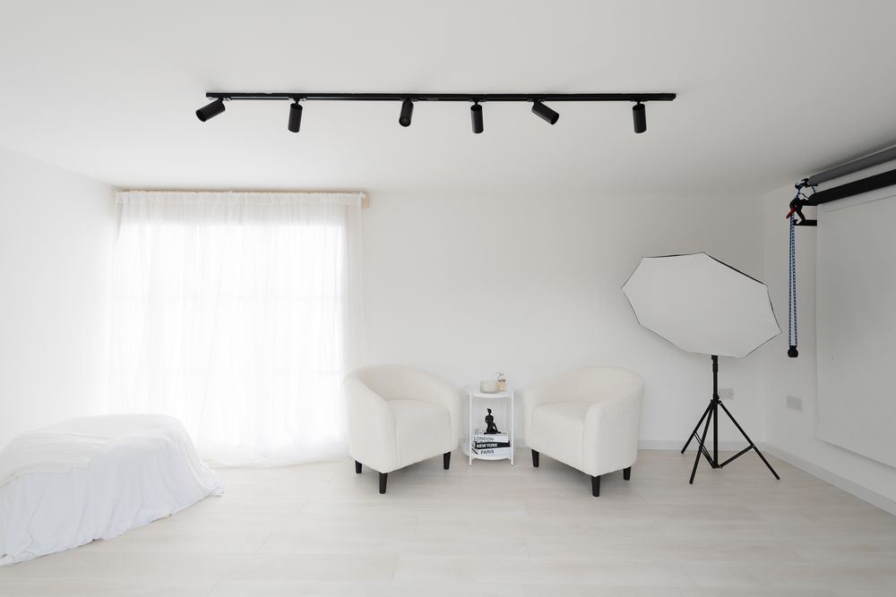 Clean white photography studio with track lighting, two chairs, softbox light setup, and sheer window curtains.