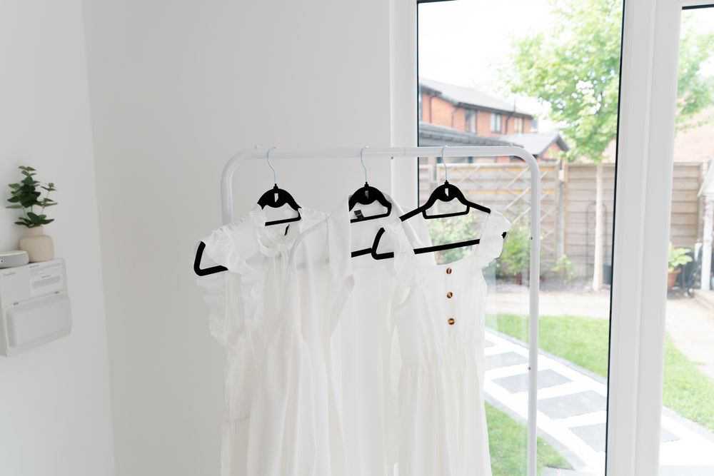 Three white wedding dresses hanging on hangers against a white wall with a window view of outdoor greenery.