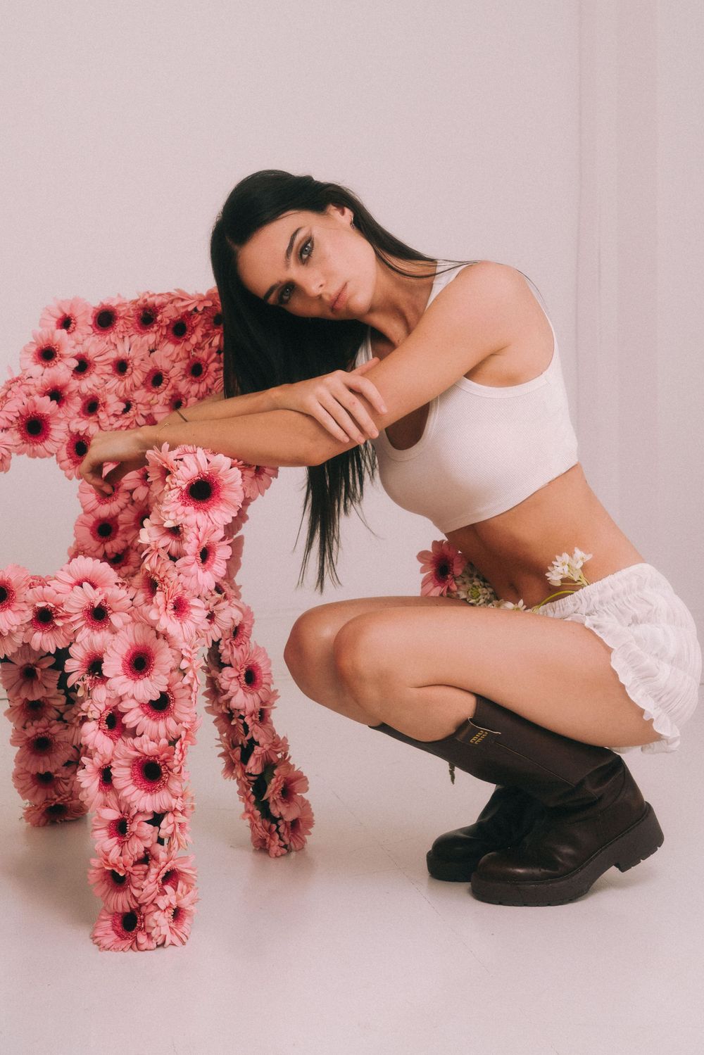 Fashion model poses with pink flower chair in white crop top and skirt with black boots against pink backdrop.