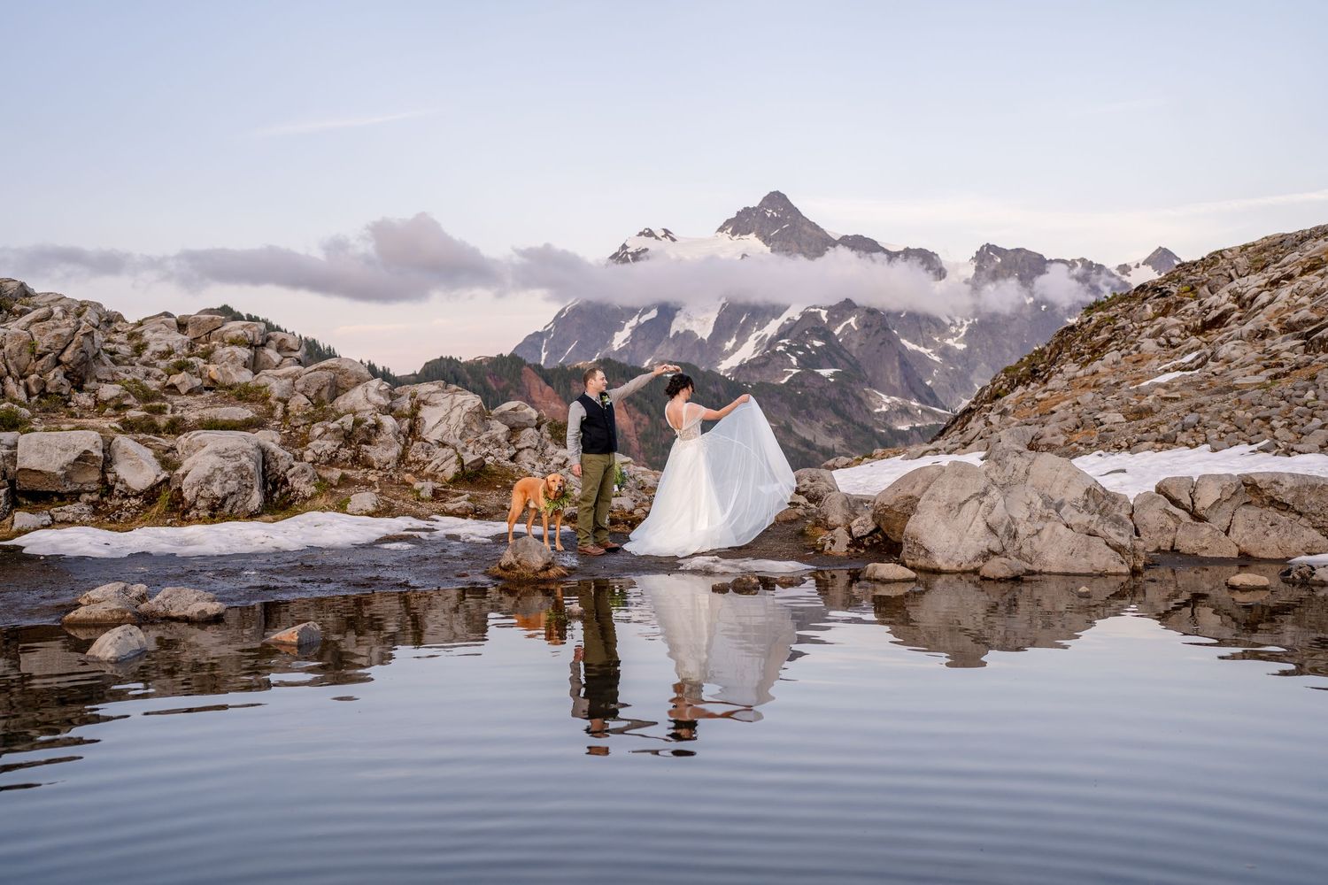 Wedding couple reflected in an alpine lake surrounded by snow-capped mountains at sunset.