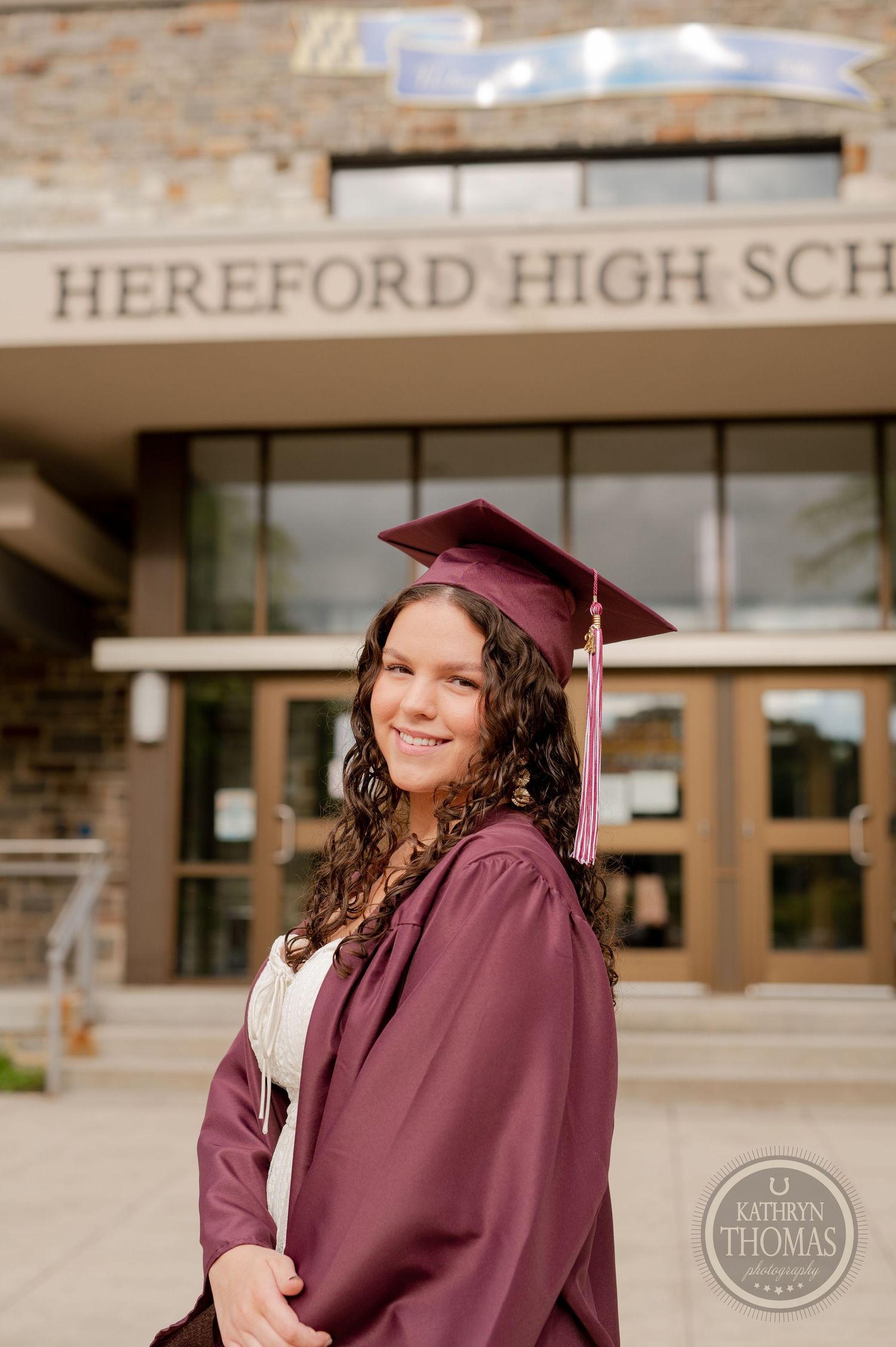 Graduate in maroon cap and gown smiles in front of Hereford High School main entrance.