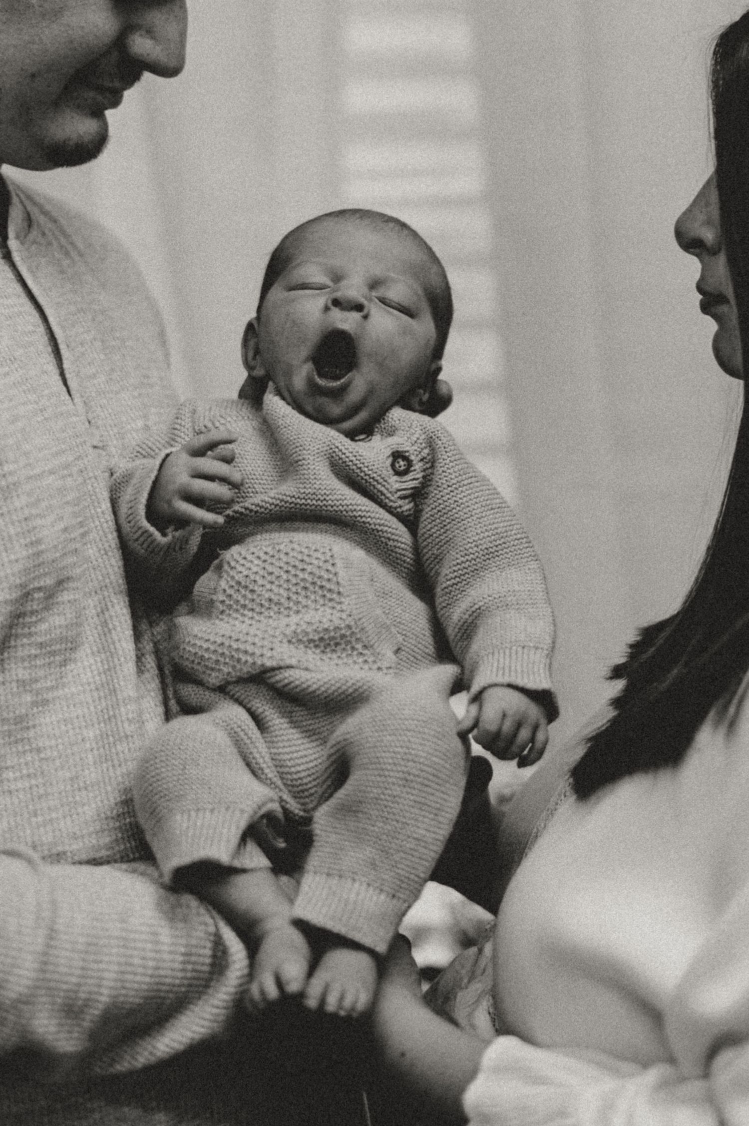 Black and white photograph of a yawning newborn baby wearing a knit sweater set held between two adults.