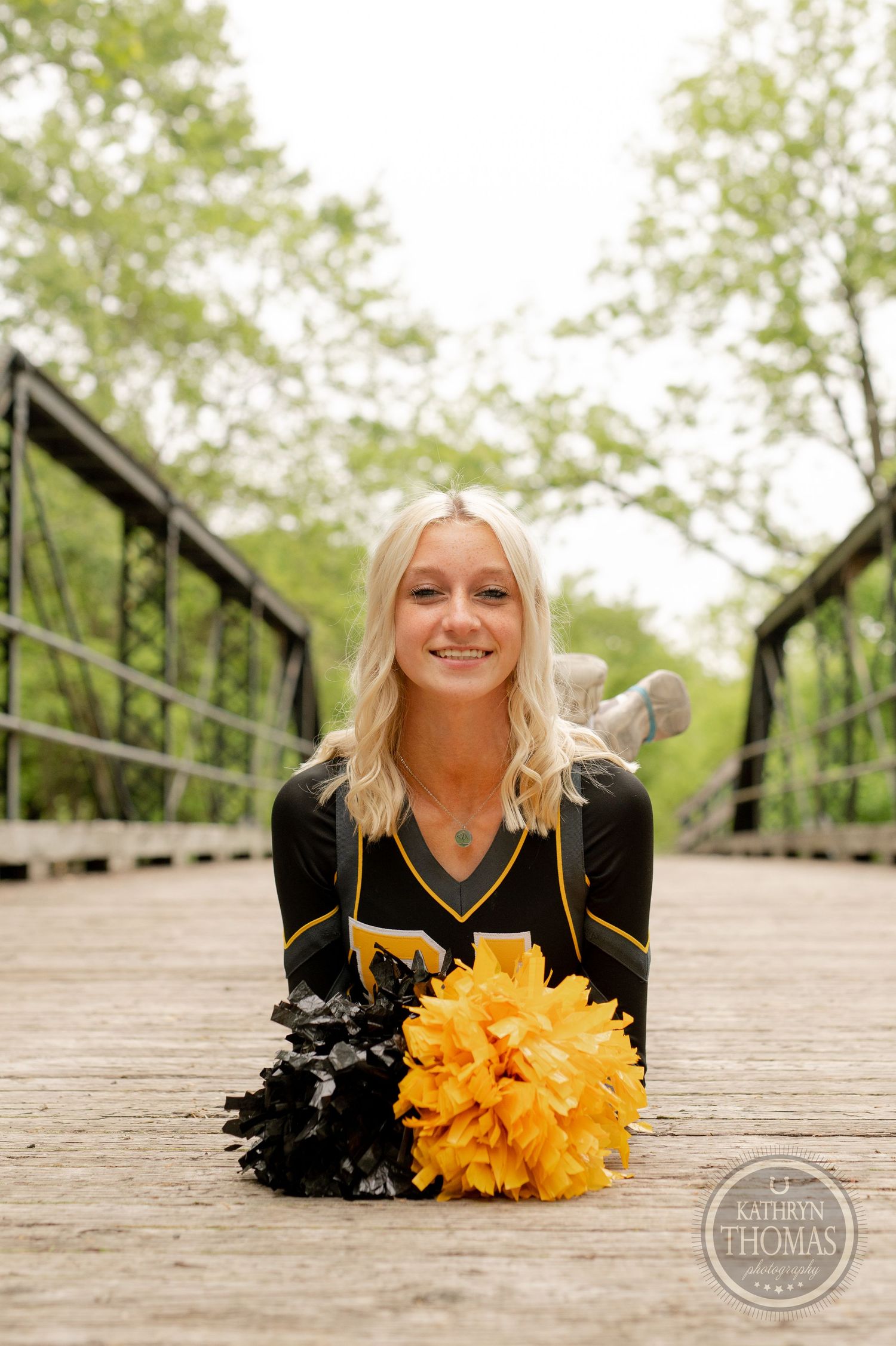 Cheerleader in black and gold uniform poses with pom poms on wooden bridge surrounded by green trees.