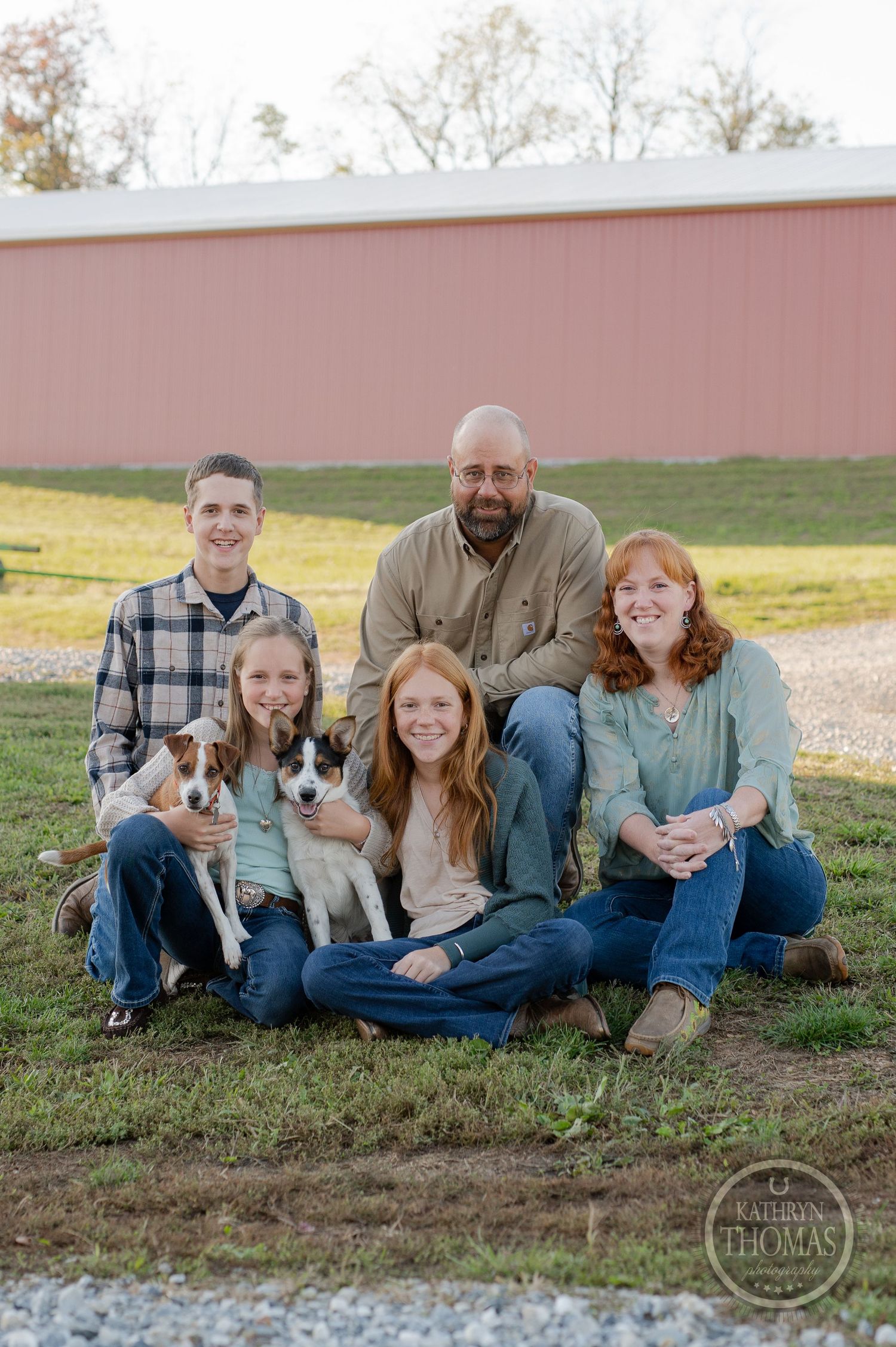 Family sits together with their two dogs in front of a pink barn.
