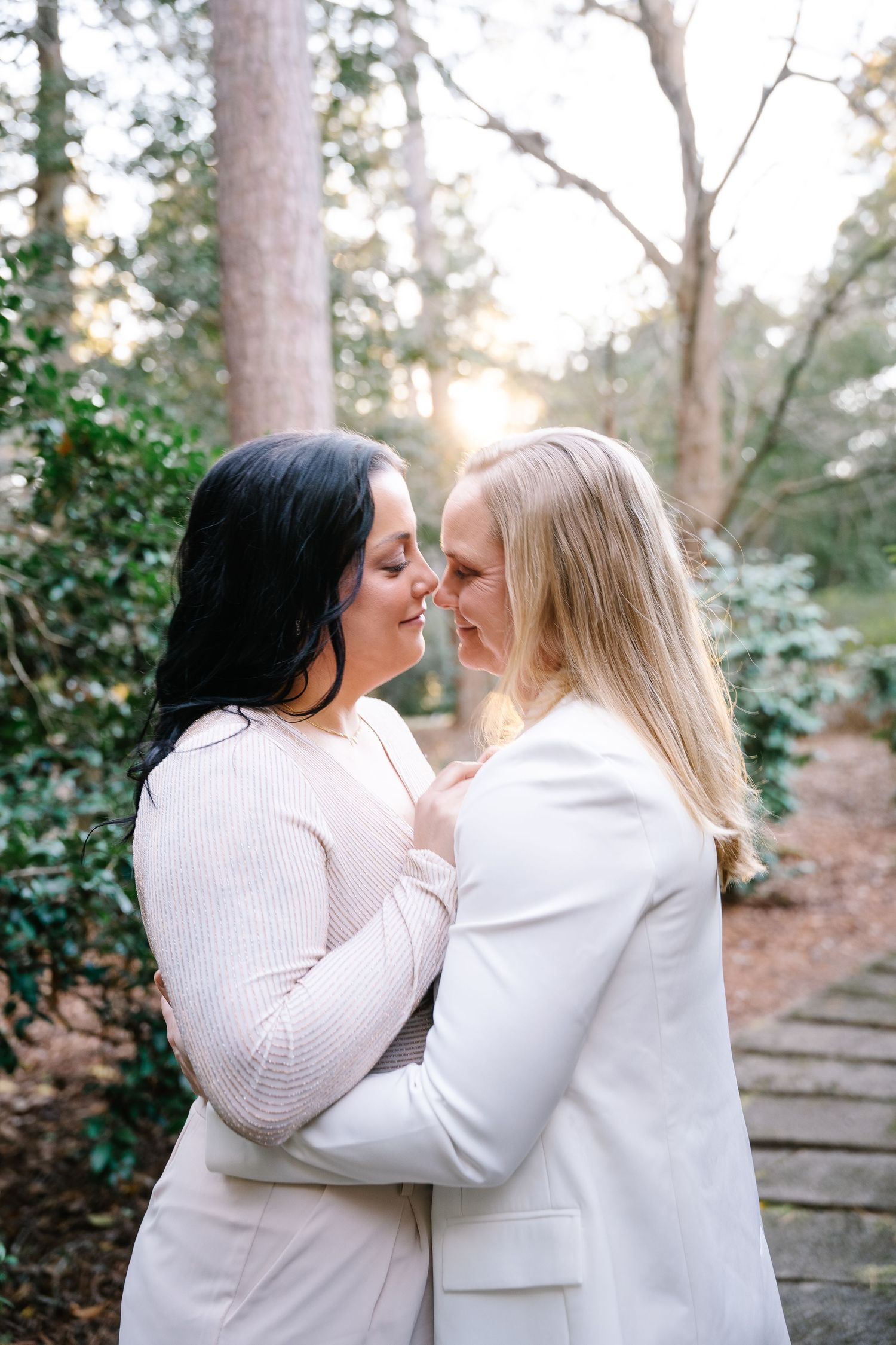 Brides share a tender moment against beautiful green trees and the setting sun during an outdoor wedding at Weymouth House in Southern Pines