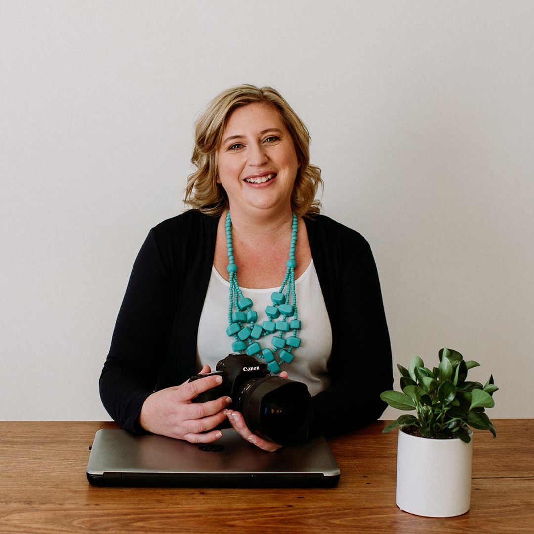 Professional photographer in black cardigan and turquoise necklace smiles while holding camera at desk.