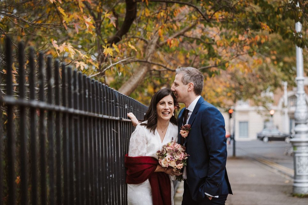 Bride and groom share a romantic moment by an iron fence amid autumn foliage, bride holding a burgundy bouquet.