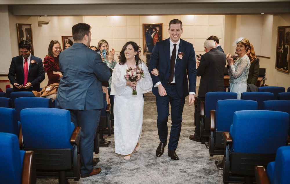 Joyful newlyweds walk down the aisle at a civil wedding ceremony, bride holding a bouquet, guests applauding in blue chairs.