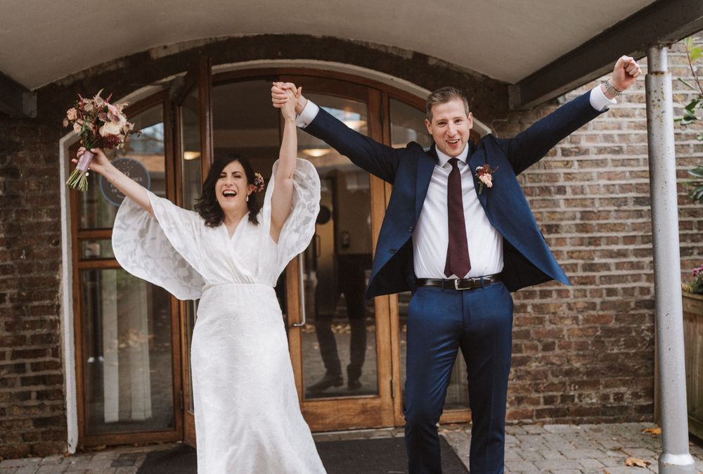 Joyful bride and groom raising hands outside brick building after wedding ceremony.
