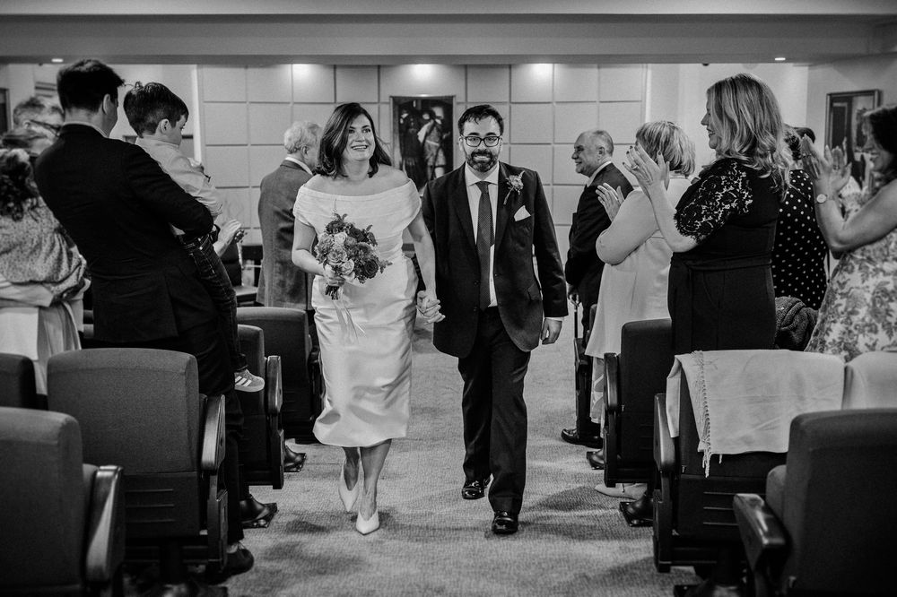 Black and white photo of a smiling couple walking down the aisle after their wedding ceremony, guests applauding.