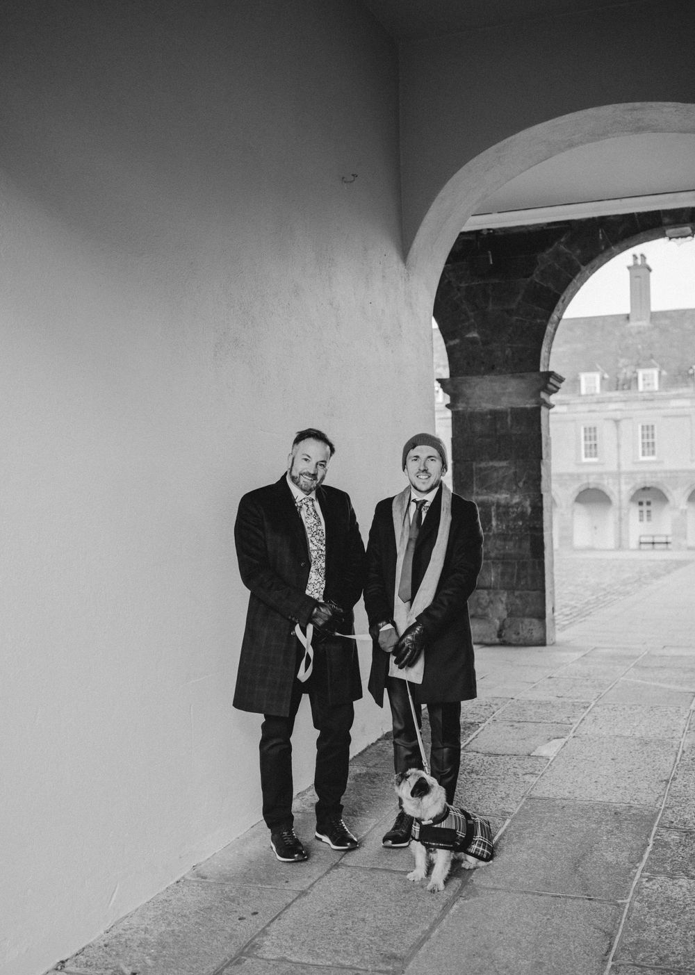 Two people in formal coats pose with a small dog under a stone archway.