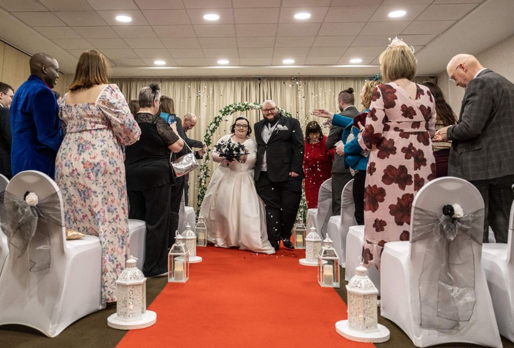 Wedding ceremony guests stand along sides of red-carpeted aisle with white covered chairs decorated with sashes in banquet hall.