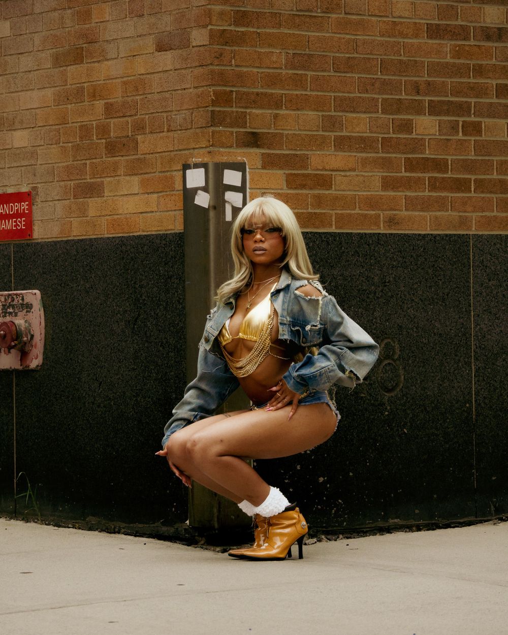 Person wearing a blonde wig, gold bodysuit and denim jacket poses crouching against brick wall in platform gold shoes.