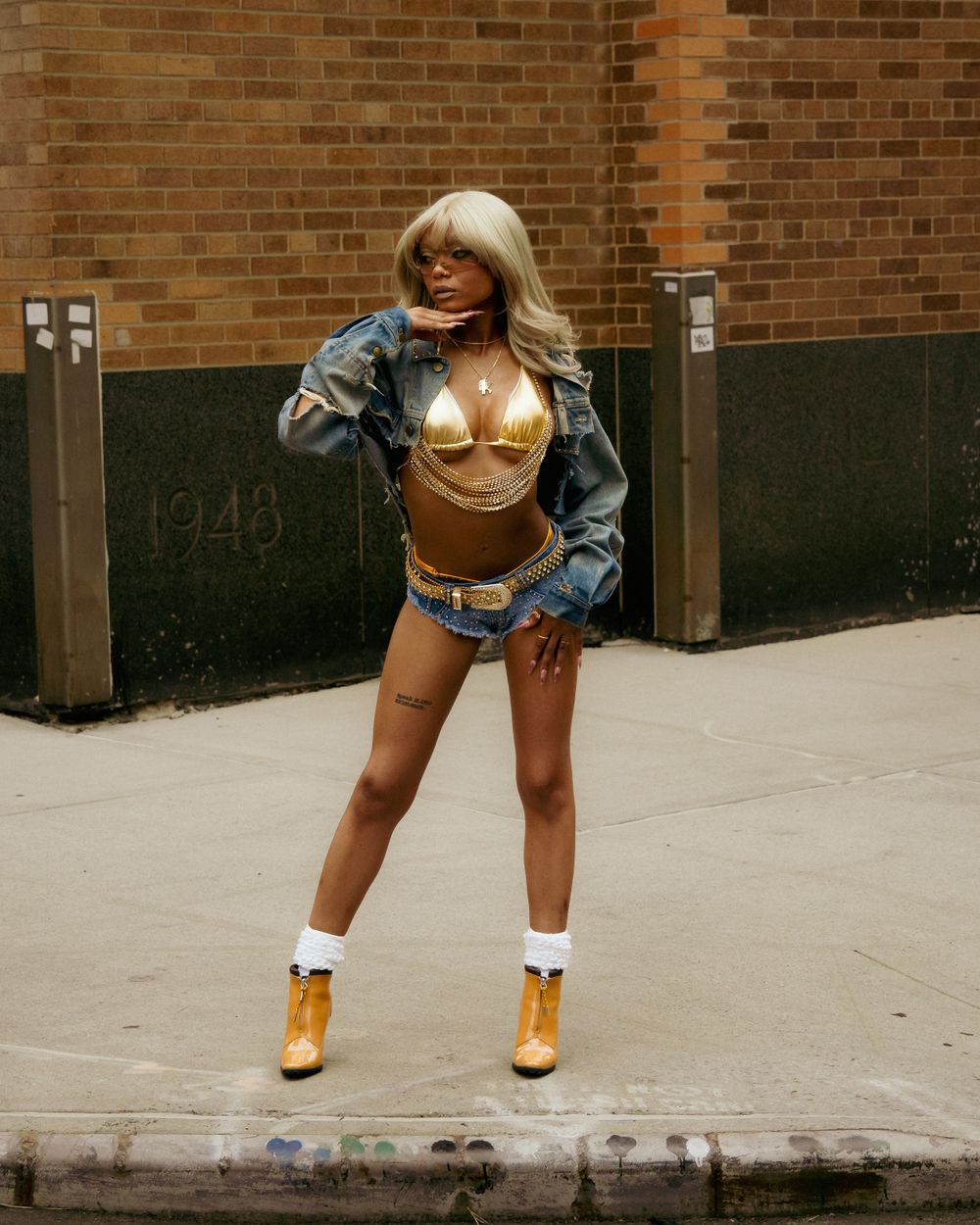 Fashionable model in denim jacket and shorts poses against brick wall wearing gold boots and white socks.