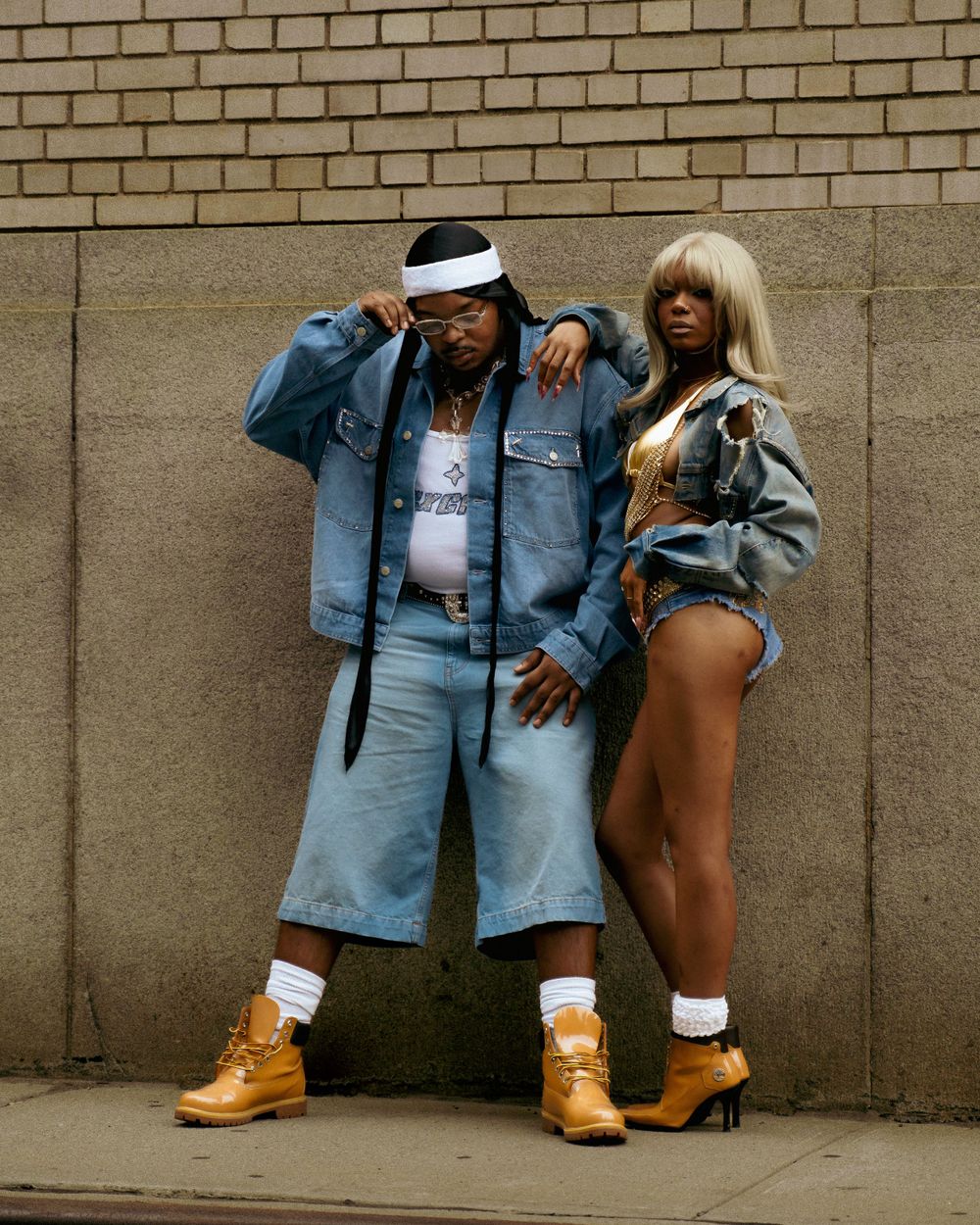 Two people pose stylishly against a brick wall wearing denim outfits, sunglasses, and brown boots.