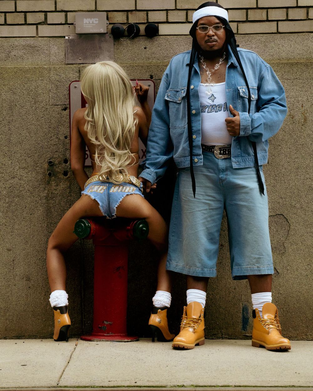 Two people wearing matching denim outfits and tan boots pose against a brick wall with a red fire hydrant.