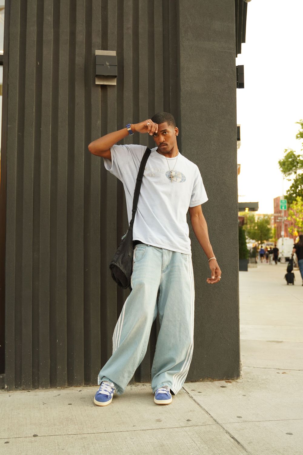 Urban street fashion look with white t-shirt, light baggy jeans, black crossbody bag and blue sneakers against city wall.
