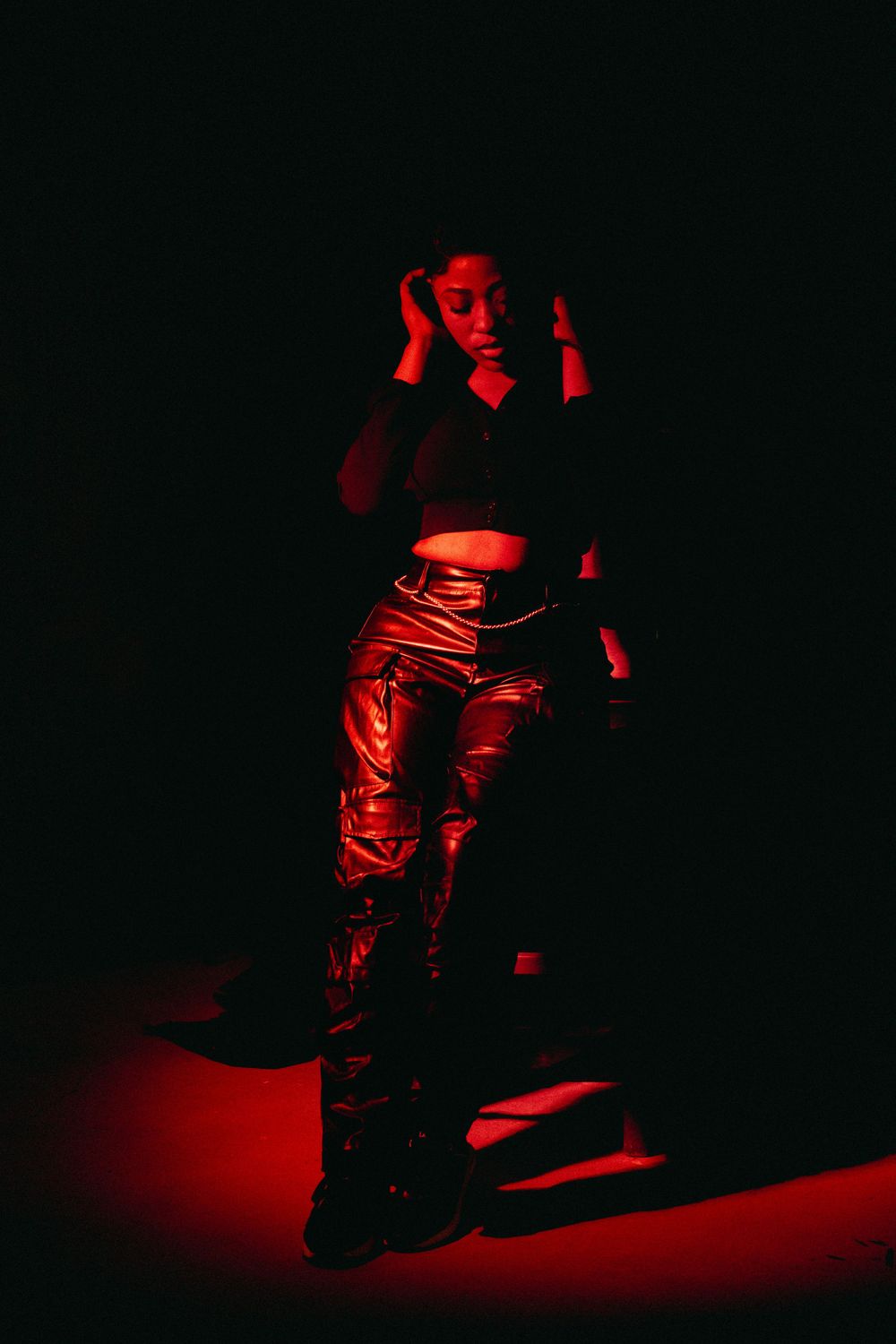 Dramatically lit figure in black clothing poses against dark background with red lighting effects.