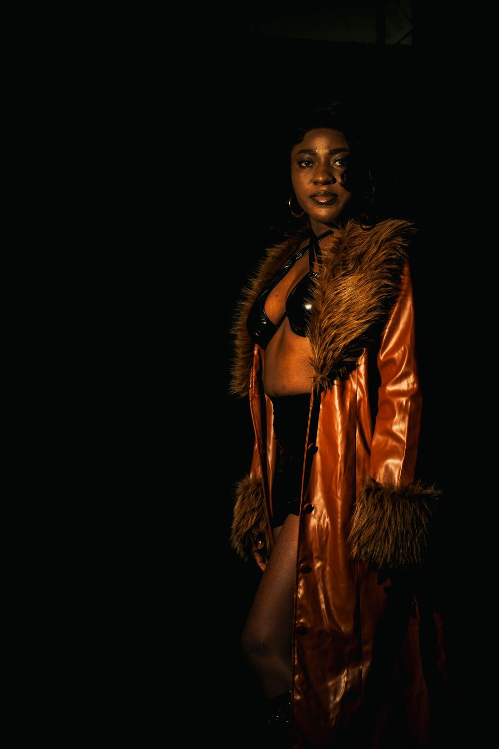 A person poses in a dramatic brown leather coat with fur trim against a dark background in moody lighting.