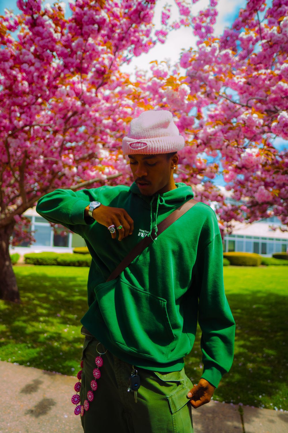 Person in bright green hoodie posing against vibrant pink cherry blossoms on a sunny spring day.
