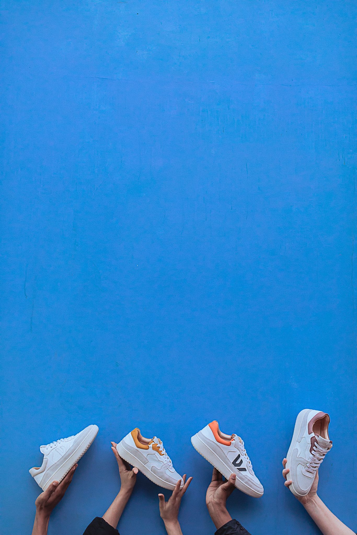 Hands holding up multiple pairs of white sneakers against bright blue background.