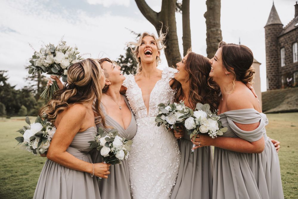 Wedding party shares joyful candid moment with bridesmaids in gray dresses holding white bouquets at Meldrum House Hotel Aberdeenshire.