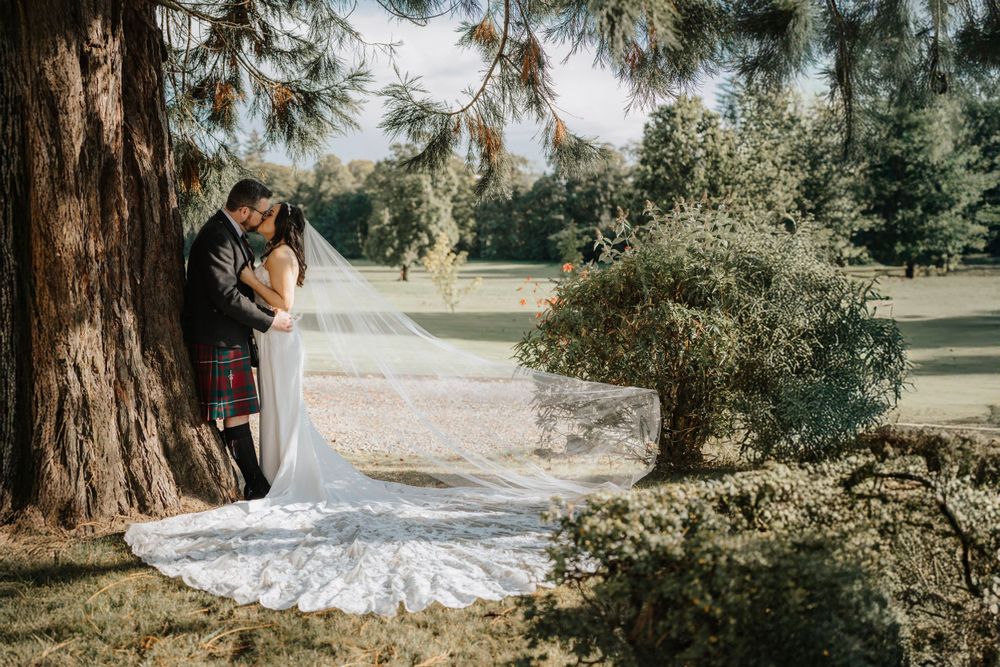 Wedding couple poses intimately by large tree trunk on manicured grounds at Thainstone House Hotel Aberdeenshire.