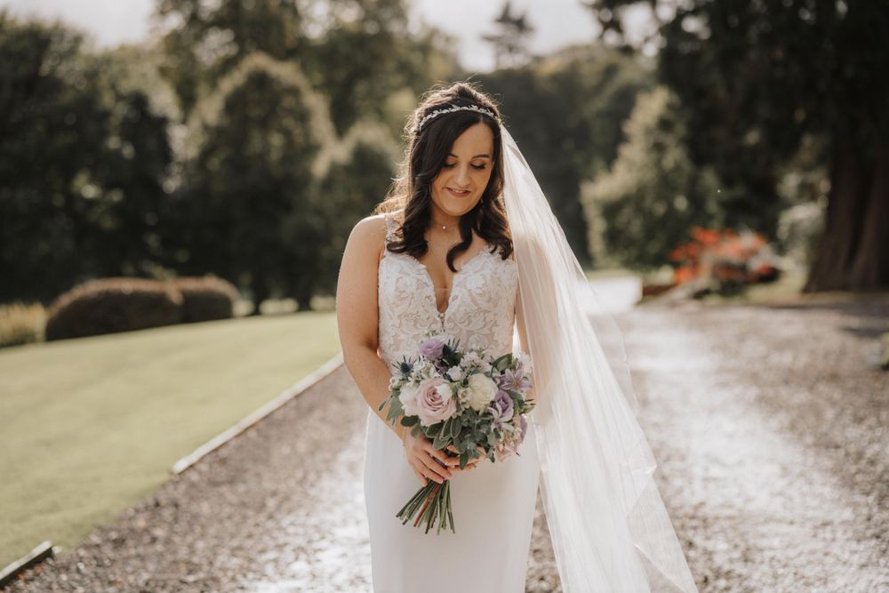 Bride in fitted white dress holds purple and white bouquet on gravel path surrounded by greenery at Thainstone House Hotel Aberdeenshire.
