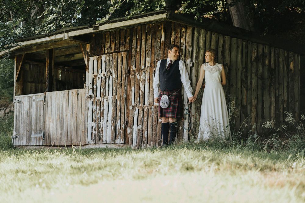 A couple stands in traditional wedding attire next to a rustic wooden barn in a countryside setting in Aberdeenshire.