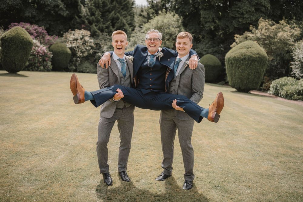 Three groomsmen in matching gray suits playfully pose with their dad on a garden lawn at elsick house Aberdeenshire.