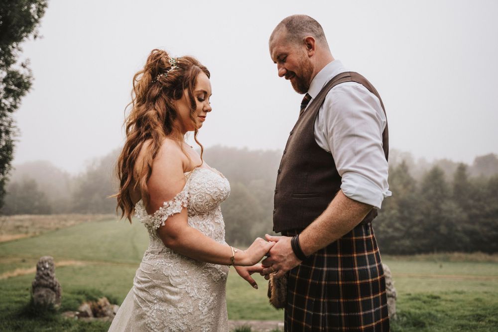 Bride in lace gown and groom in traditional kilt hold hands during misty outdoor wedding ceremony at Ardoe house Aberdeenshire.